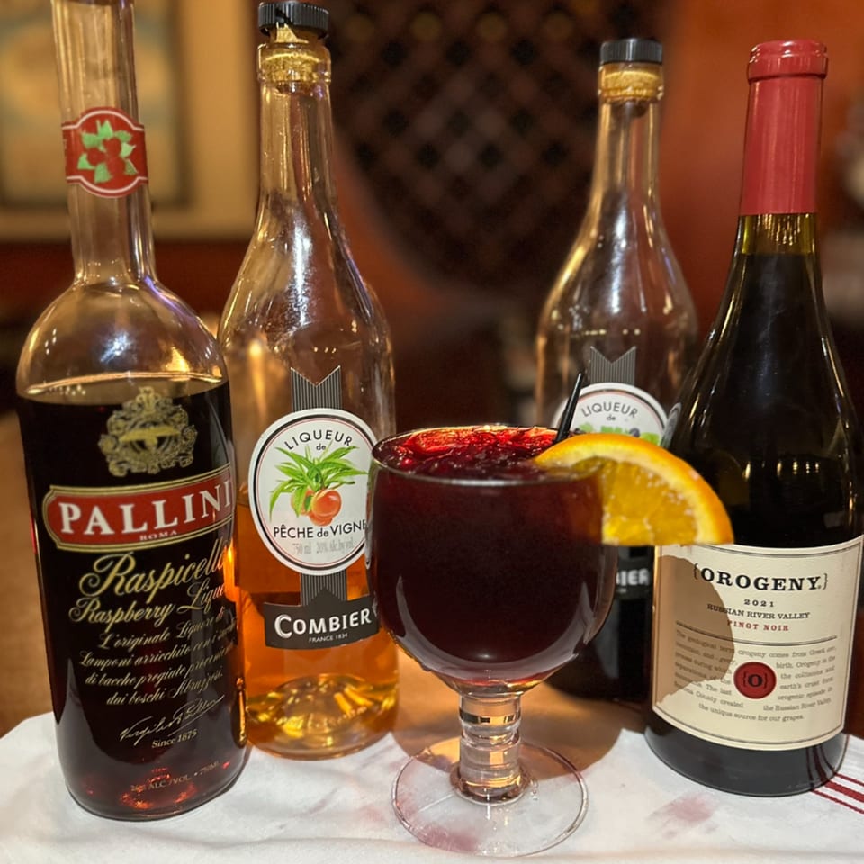 A glass of red sangria with an orange slice, surrounded by bottles of liqueur and wine.