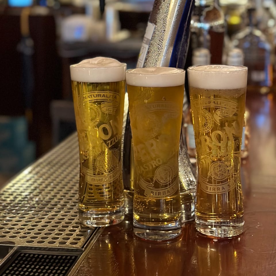 Three tall glasses of Italian Peroni beer with foamy heads sit on a bar counter.