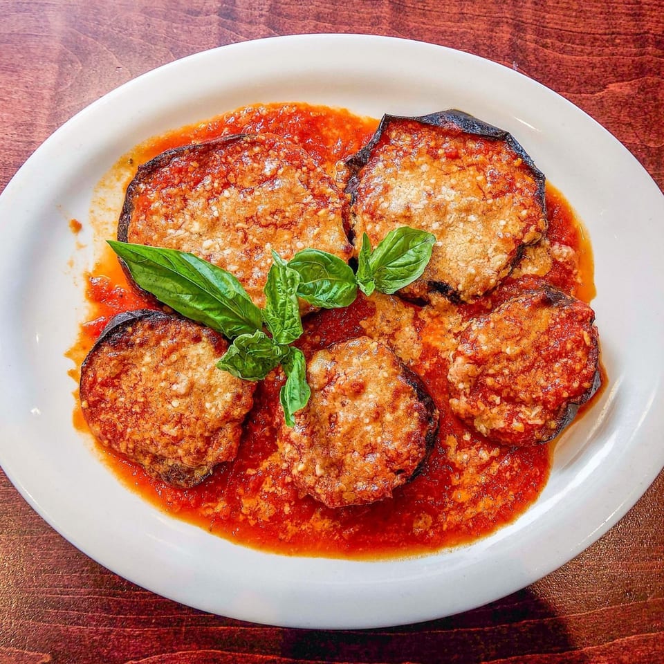 Nick's Italian eggplant parmesan, five slices covered in red sauce, cheese, and basil on a white plate.