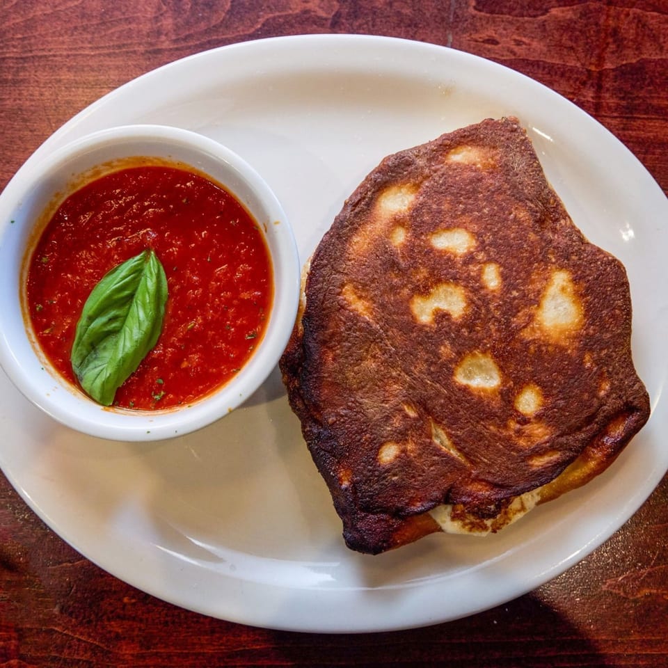 A golden-brown calzone with a side of red marinara sauce garnished with a fresh basil leaf.