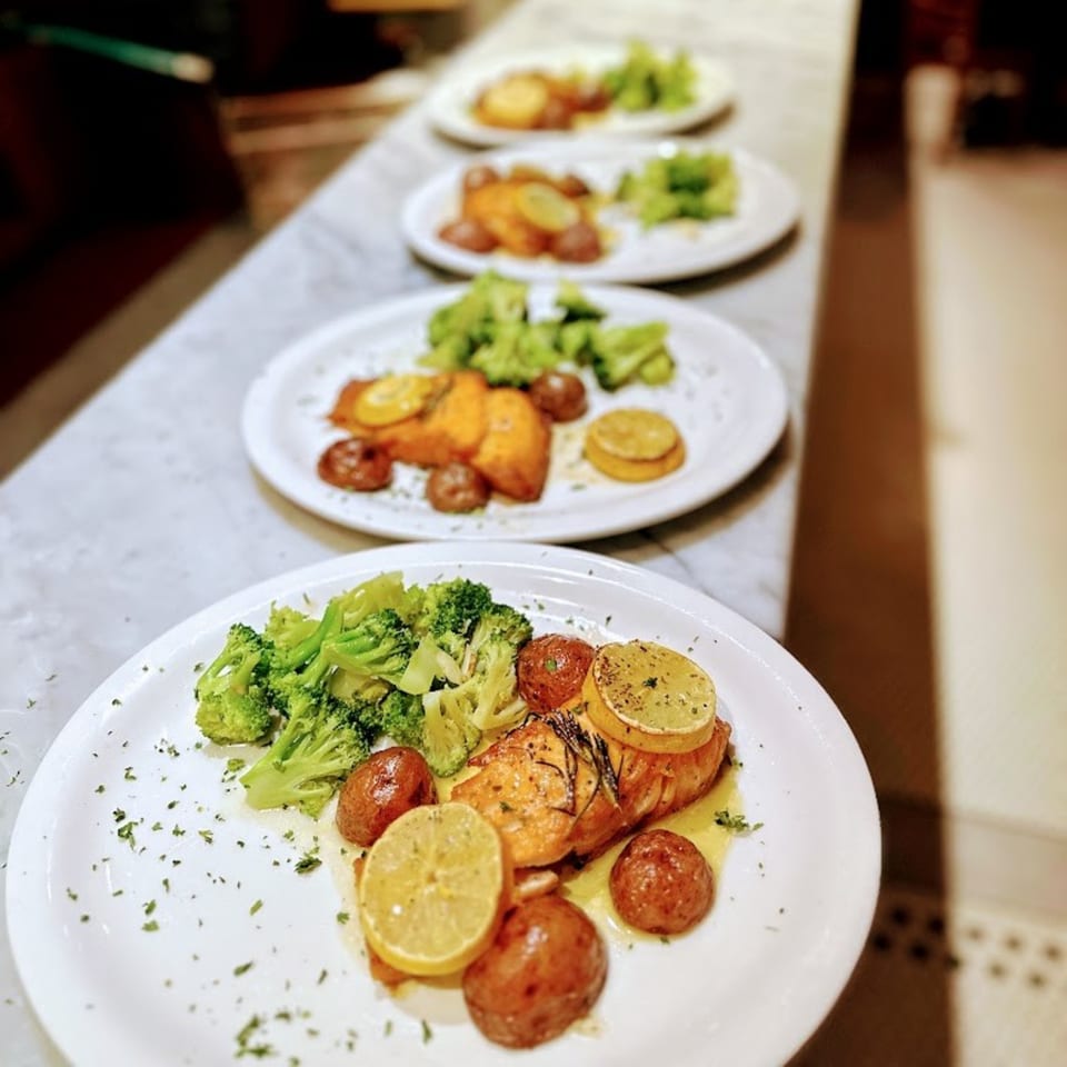 Four plates of salmon with roasted potatoes, broccoli, and lemon slices lined up on a counter.