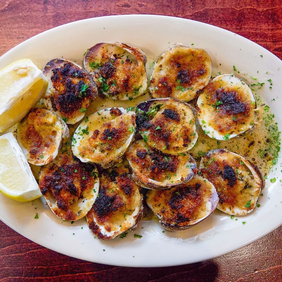 A white oval plate filled with baked clams casino, garnished with parsley and lemon wedges.