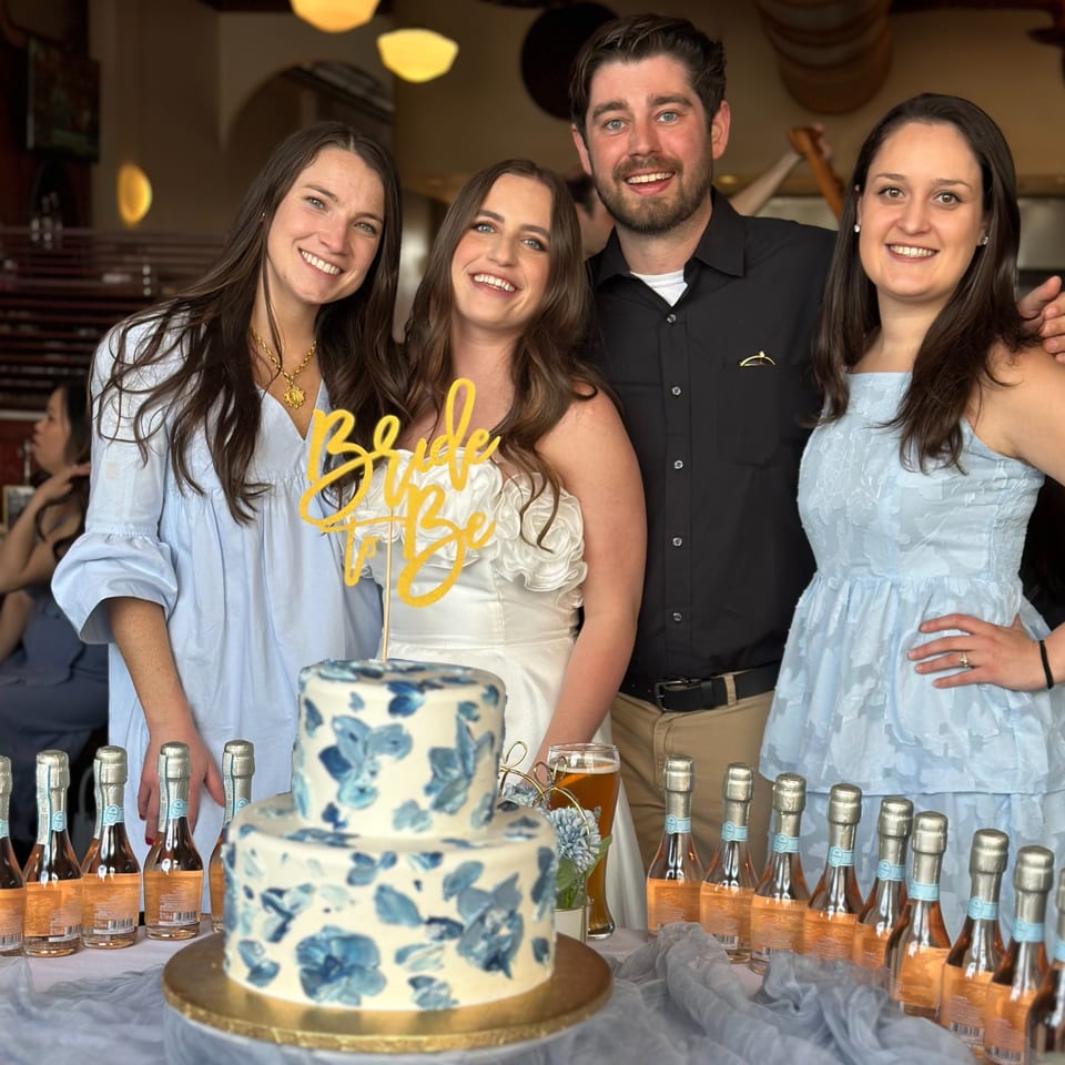 Four smiling people, including a bride-to-be, stand behind a blue floral cake and mini champagne bottles.