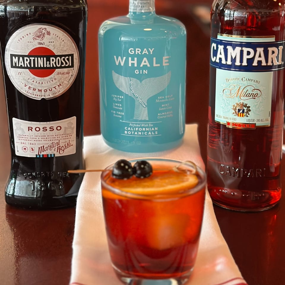 A Negroni cocktail with orange and cherries, flanked by bottles of Martini & Rossi, Gray Whale Gin, and Campari.