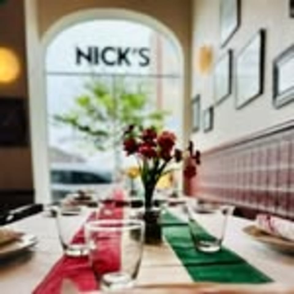 Set table with red and green runners, glasses, and a flower vase, with "NICK'S" visible through a window.